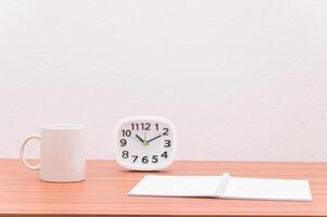 A coffee mug, notebook and a clock on a wooden table photo