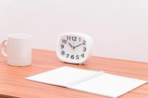 A white mug, notebook and a clock on a wooden table photo