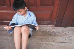 Niño sentado contra la puerta de madera leyendo un libro sobre un piso de madera foto