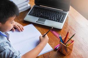 Niño haciendo los deberes con el cuaderno, la computadora portátil, la grapadora y una taza de lápices en un escritorio de madera foto