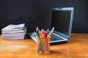 Cup of pencils, a laptop, and a stack of books on a wooden table next to a black wall photo