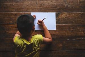 Niño visto desde atrás escribiendo en papel sobre un piso de madera foto