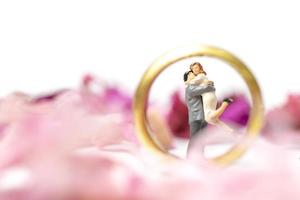 Pareja en miniatura abrazándose con un anillo de bodas y pétalos de rosa aislado sobre un fondo blanco. foto