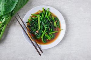 Fried Chinese kale with oyster sauce with chopsticks on a white plate next to fresh Chinese kale on wooden table photo