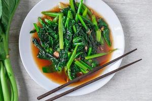 Fried Chinese kale with oyster sauce with chopsticks next to fresh Chinese kale on a white plate on wooden table photo