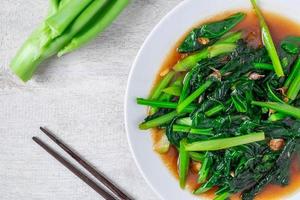 Fried Chinese kale with oyster sauce with chopsticks next to fresh Chinese kale on a white plate on wooden table photo