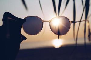 Mujer sostiene gafas de sol en la playa al atardecer de fondo de verano foto