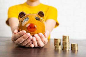 Close-up of woman in yellow shirt holding piggy bank next to stacks of coins
