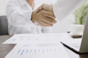 Business people shake hands next to laptop with papers of charts and graphs