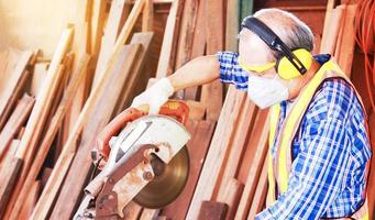 Elderly Asian carpenter craftsman uses circular saw to process wood for furniture