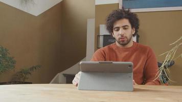 joven de Oriente Medio se sienta a la mesa, mira la computadora portátil frente a él, habla y escucha