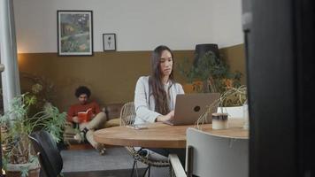 Joven mujer de raza mixta en la mesa trabajando en la computadora portátil, hablando, sonriendo, joven de Oriente Medio en el sofá, libro de lectura, riendo