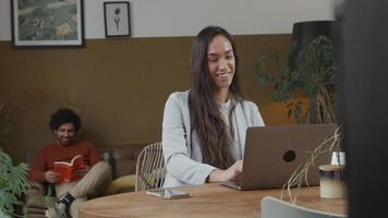 Young mixed race woman at table working on laptop, talking, smiling, young Middle Eastern man on sofa, reading book, laughing video