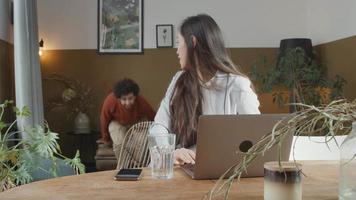 Joven mujer de raza mixta se sienta a la mesa con un portátil, se vuelve hacia un joven de Oriente Medio en el sofá, el hombre se une a la mujer, ambos saludando a la pantalla video