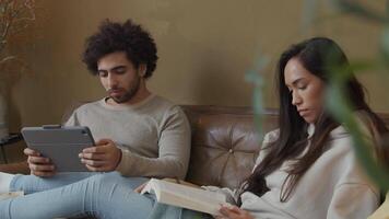 Young mixed race woman and young Middle Eastern man sitting on couch, woman reads book, man looks at tablet video