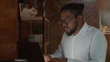 hombre negro con gafas, sentado a la mesa, escribiendo en la computadora portátil video