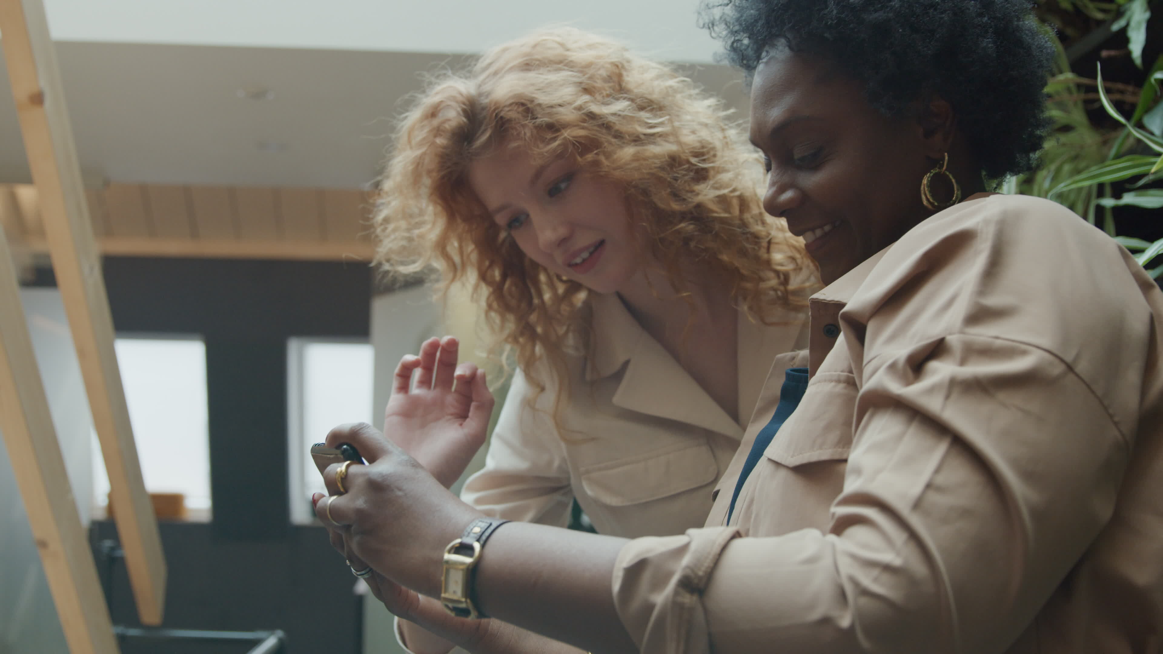 Black mature woman and young white woman standing in hallway, looking and pointing at mobile phone, smiling