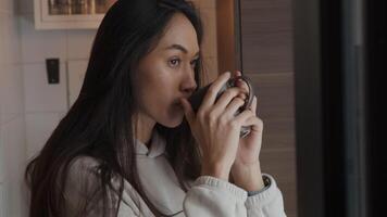 Head shot of young mixed race woman, holding cup with both hands, taking a sip, looking through window contemplatively, holding cup, shot in mirror, partly blurred on the right side video