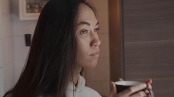 Jeune métisse femme marche tenant tasse à deux mains, se dirige vers la fenêtre, en prenant une gorgée, regardant à travers la fenêtre de manière contemplative video