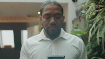 Black man stands in hallway, looking at mobile phone holding in front of him, zoom, looks into camera and smiles video