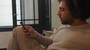 junger Mann aus dem Nahen Osten mit Kopfhörer auf dem Kopf, sitzt auf dem Sofa, berührt den Bildschirm des Mobiltelefons, bewegt den Kopf und singt ein bisschen