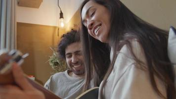 Young mixed race woman and young Middle Eastern man sitting on couch, woman plays guitar whilst talking and laughing with man video