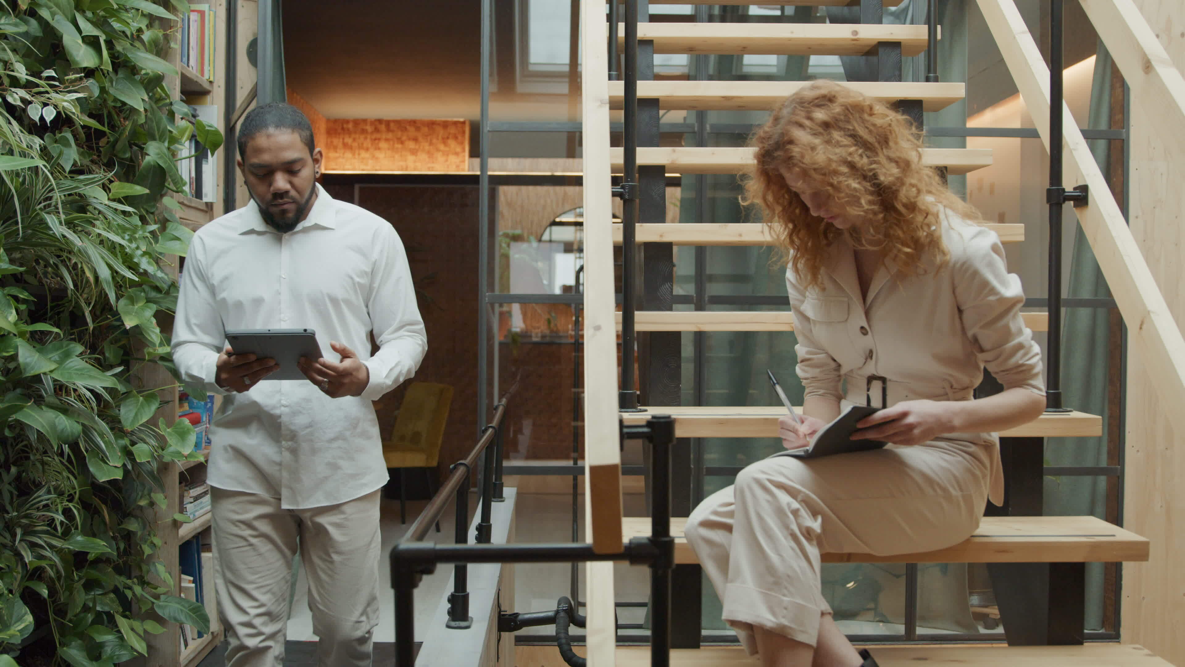 Black man walks with tablet shows it to young white woman who sits on the stairs