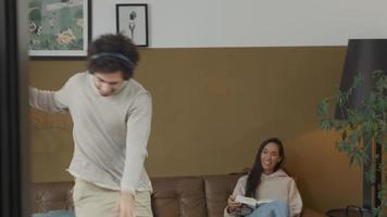 giovane uomo mediorientale con le cuffie balla espressamente sulla musica in salotto, la giovane donna di razza mista sul divano ride video