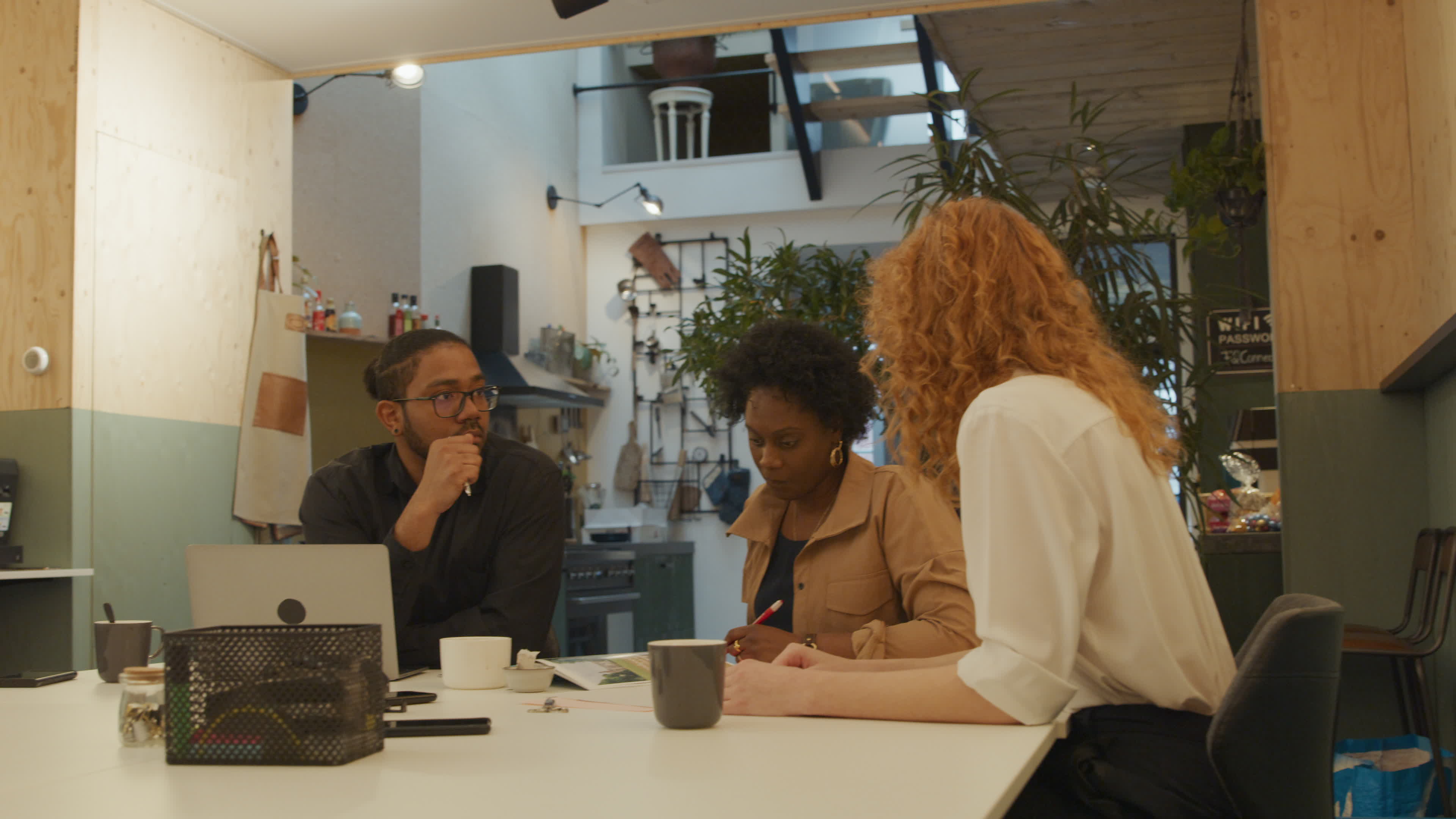Black mature woman, white young woman and black man wearing glasses, sitting at table in office, talking, white woman gestures