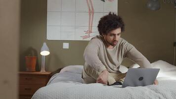 Young Middle Eastern man sits on bed, crossed legs, laptop in front of him, talking to screen, blurred doorframe on the left side video