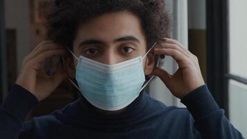 Close up and slow motion of young Middle Eastern man, looking seriously into camera lens, putting on face mask with both hands, keeps looking into camera video