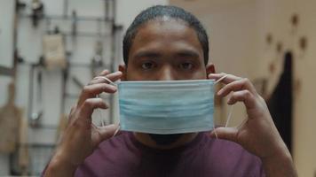 Close up of black man, looking down, lifts head, looking seriously into camera lens, puts on face mask with both hands, adjusting mask video