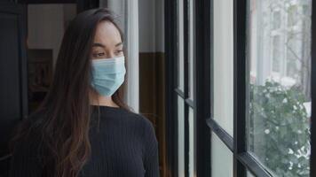 Medium shot and slow motion of young mixed race woman with face mask on, next to window, looks through window, turns slightly to window, eyelashes, blinking video