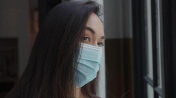 Cerca de joven mujer de raza mixta con mascarilla, junto a la ventana, mirando a través de la ventana video