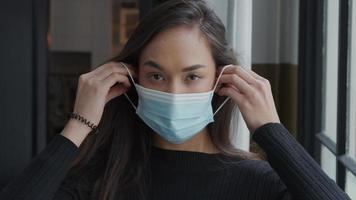 Head shot of young mixed race woman looking down, lifts head and hands, holding face mask, puts on face mask, adjusts it, looks in lens video