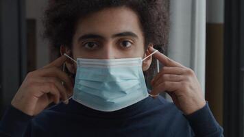 Close up and slow motion of young Middle Eastern man, looking seriously into camera lens, putting on face mask with both hands, adjusting mask, keeps looking into camera video