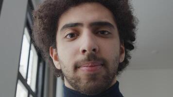 Close up of young Middle Eastern man, head slightly turned down, looking, smiling a bit, into camera lens, eyes following video