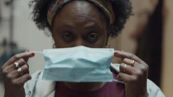 Close up and slow motion of black mature woman, smiles and frowns, looks into camera, puts on face mask with both hands, adjusting it, keeps looking into camera lens video