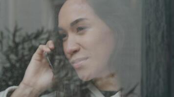 Close up made outside of young Middle Eastern man looking at and calling with, camera moves, young mixed race woman who stands inside with mobile phone on ear looking at man video