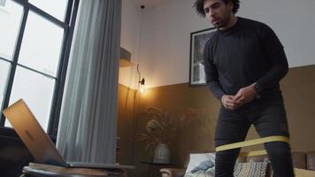 jonge man uit het Midden-Oosten in de woonkamer, staand, vooraan kijkend naar laptop, weerstandsband boven de knieën, zijwaarts stappen met gebogen knieën