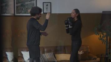 Young Middle Eastern man stands in living room, hands in front of him, catching punches of young mixed race woman with boxing gloves