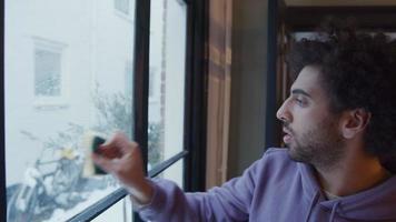 Young Middle Eastern man cleans window with sponge and tries to dry window with squeegee, man laughs
