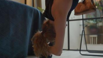 jovem mulher branca em pé na sala de estar, movendo os braços e as costas, flexionando e alongando, pernas montadas, fazendo aulas de ginástica online video