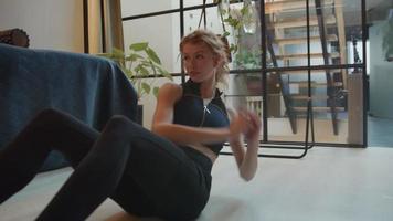 Giovane donna bianca è seduta in soggiorno sul pavimento, ginocchia piegate, schiena sollevata, mani giunte, braccia che si muovono lateralmente, lezione di ginnastica online