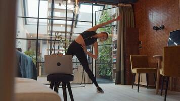 Wide shot of young white woman in living room, in straddle, moving her arms and her back in various directions, doing exercises online, laptop in front of her video
