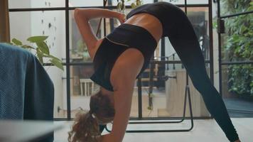 Young white woman standing in living room, moving her arms and her back, legs in straddle, doing exercise class online with laptop in front of her video