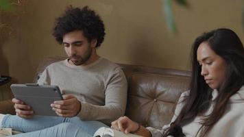 Young mixed race woman and young Middle Eastern man sitting on couch, woman reads book, man watches and touches tablet