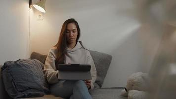 giovane donna di razza mista si siede in un angolo del soggiorno, guardando e toccando lo schermo del tablet sulla gamba video