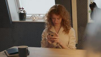 Young white woman sits at table with mobile phone, typing and having a call video