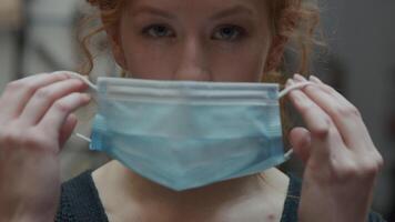 Close up of young white woman, eyes turned down, puts on face mask with both hands, adjusting mask whilst eyes looking into camera video